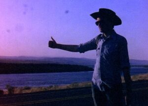 A man in a cowboy hat with his thumb out to hitch a ride in a purple-tinged light.