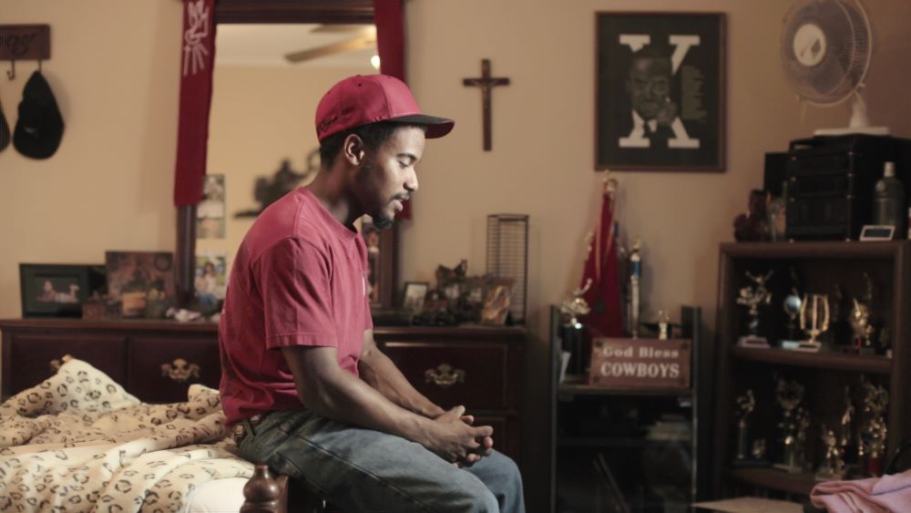 Dark-skinned man sitting on the end of a bed, in ball cap, red T-shirt and jeans, hands folded, with a cross, a God Bless Cowboys sign, mirror, trophies, dresser, fan and other items in the background