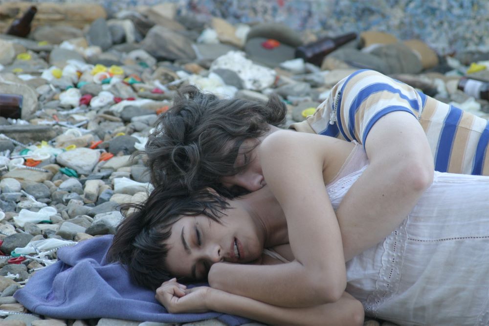 A young, dark-haired woman and a young, dark-haired man sleep, spooning, on a blanket in a littered landscape.
