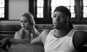Black man with tear-filled eyes sitting in what appears to be a pew, a woman with long, blond hair sitting behind him
