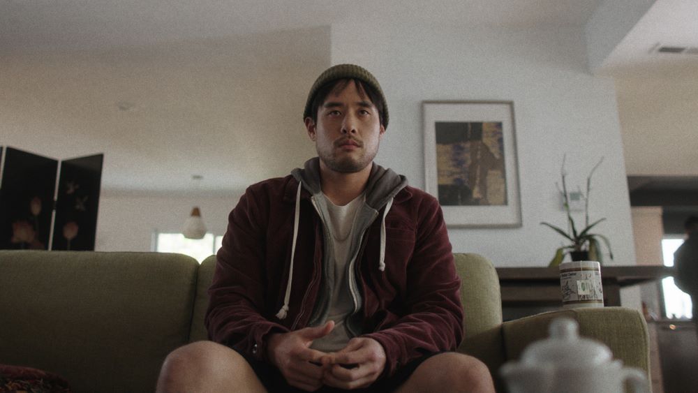 Young bearded Asian man in stocking cap, zippered hoodie, and shorts sits on a couch.