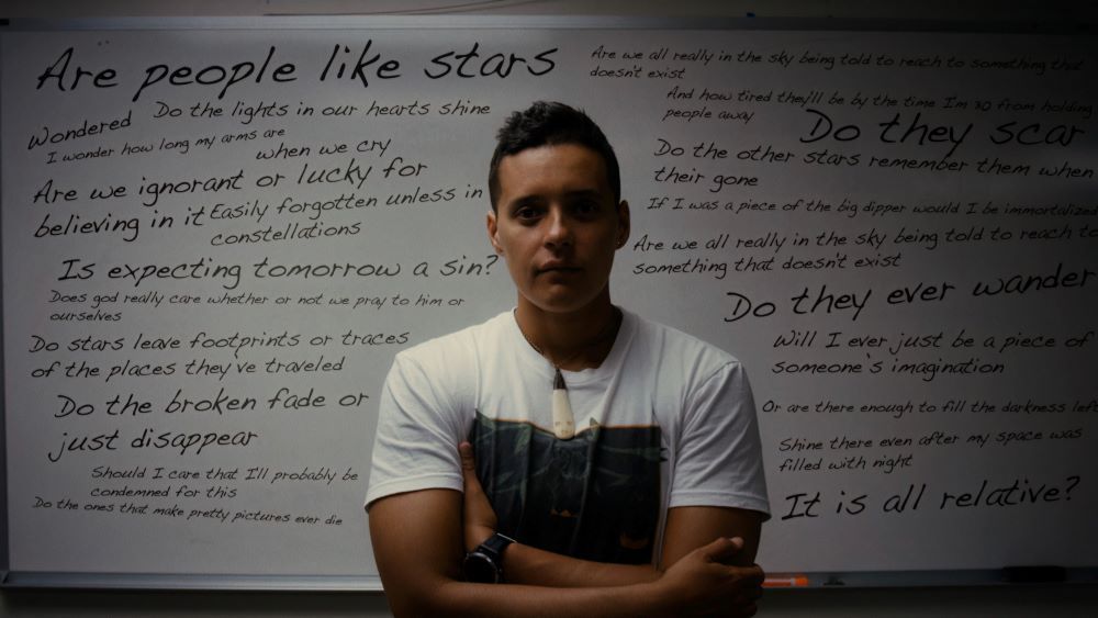 Woman in white T-shirt, arms crossed, standing in front of a white board filled with writing