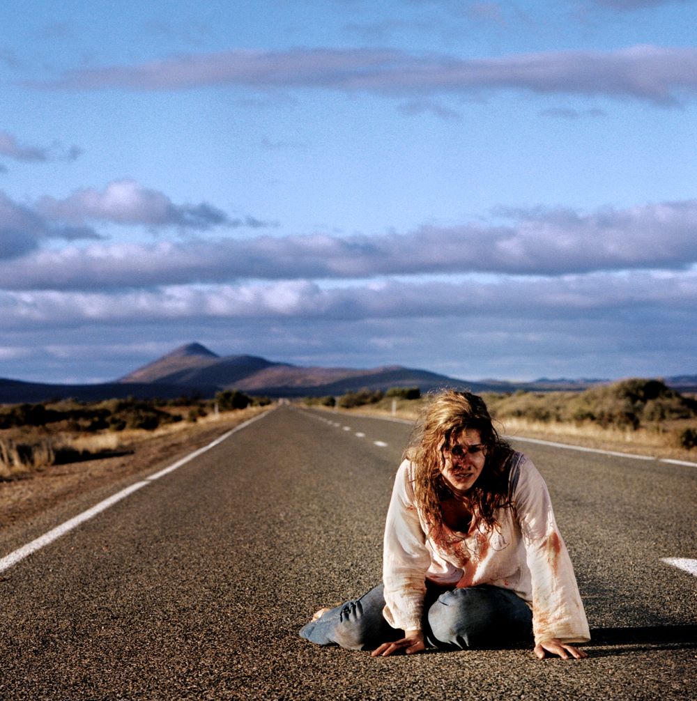 A bloodied woman with tangled long hair sits, legs folded under her, in the middle of a highway in an empty landscape, stark mountains in the background.