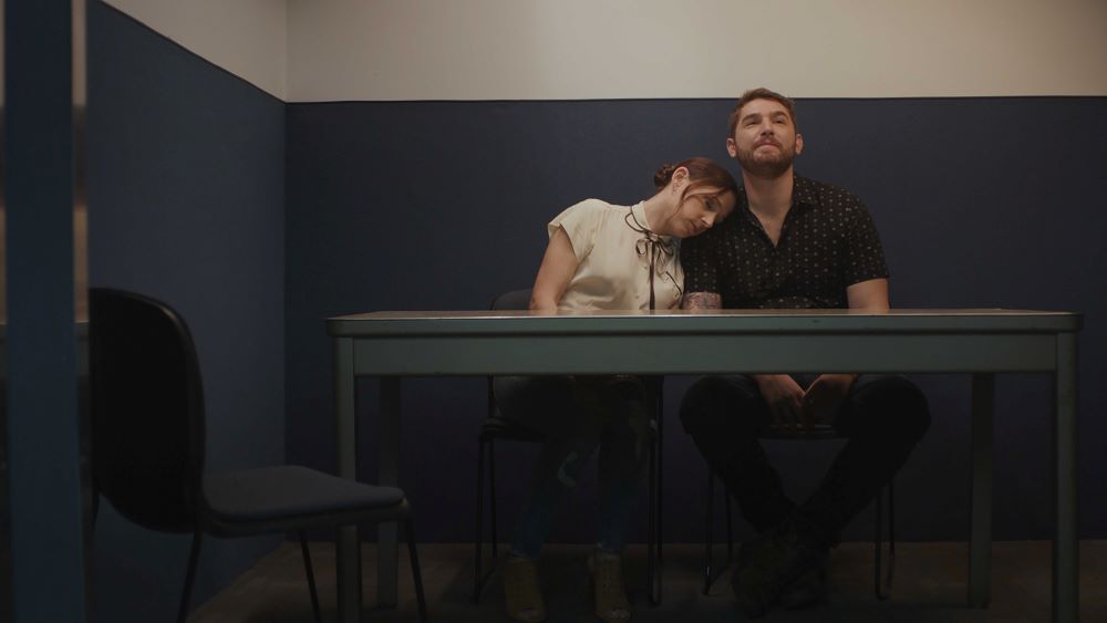 A white woman and a white man sit side-by-side at a table, the woman's head resting on the man's shoulder, the bearded man gazing upward