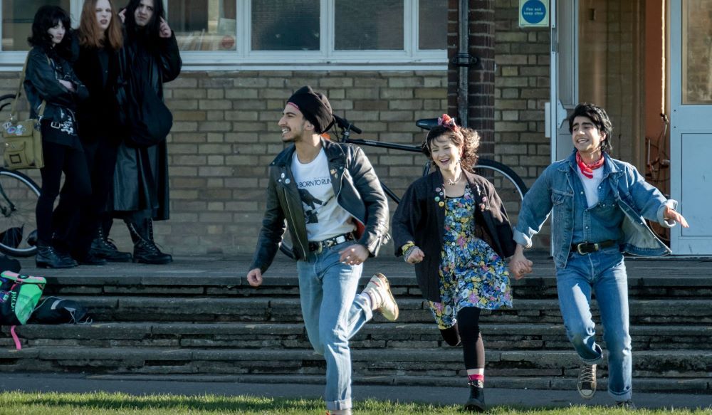 Two young men and a young woman run gleefully outside a brick building.