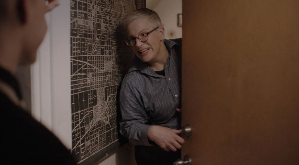 Person with short gray hair and eyeglasses, peers with an apparent smile from inside a half-opened door at an apparent visitor.