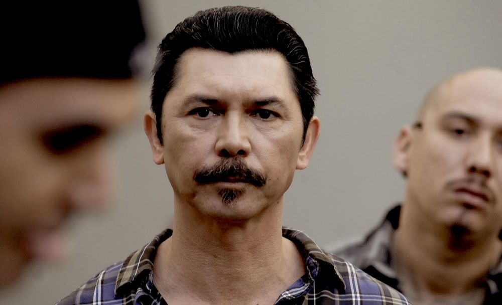 Man with dark, graying hair, mustache and soul patch, wearing a plaid shirt, stares into the camera, with blurred faces in the foreground and background.