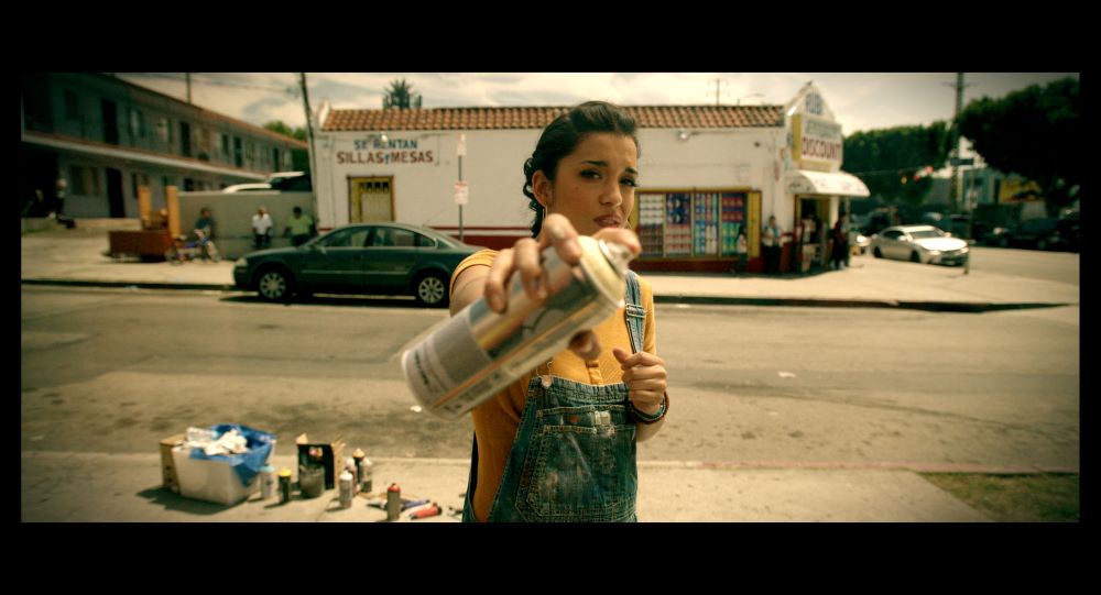 Latino woman points a spray can at the camera while standing on a sidewalk, with a container of what appears to be more spray cans and other items on the sidewalk behind her. A storefront with people standing outside, and and what appears to be a two-story motel or apartments are in the background.