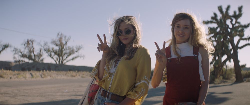 Two young blond women standing side-by-side in an outdoor setting, flashing the victory sign.