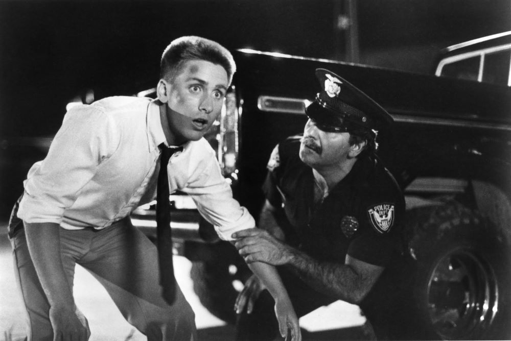 Policeman grips the arm of a young man with short hair, a bump on his forehead, wearing a light shirt and dark tie, looking with wide eyes toward something off screen.
