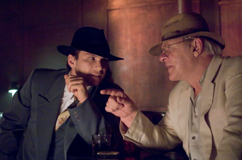 Young man in a dark suit and fedora confers with an older man with gray hair and eyeglasses in a light-colored suit and fedora