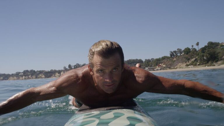 Closeup of a man lying chest-down on a surfboard in the water, arms paddling to the side