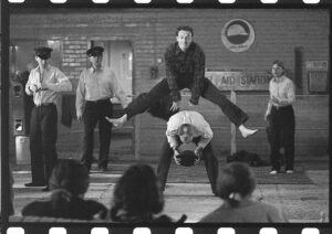 Man leap-frogs over another, with three other men, two wearing police officer hats, in the background on what appears to be a makeshift stage