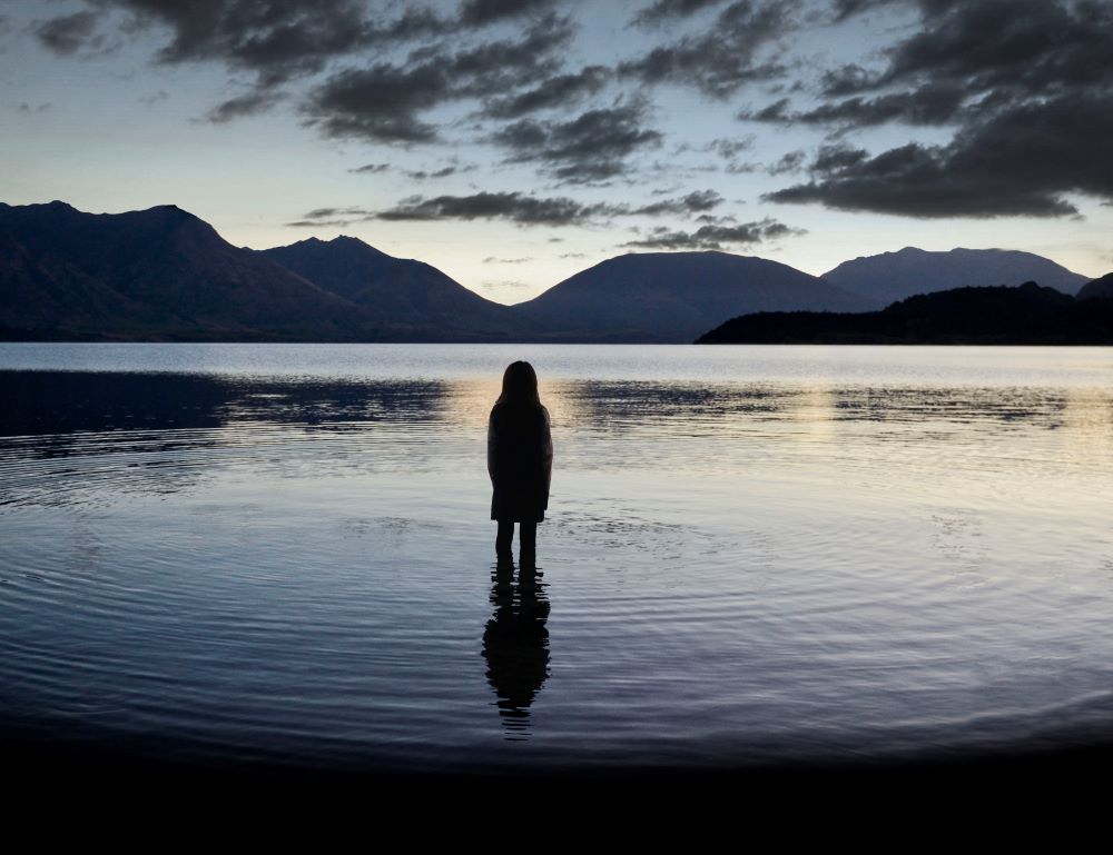 Against a backdrop of mountains and a cloudy night sky, a girl stands mid-calf in a body of water, her back to the camera.