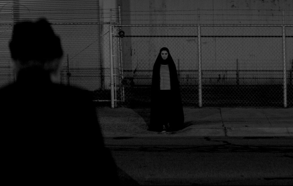 A dark-clothed figure with a still face leaning up against a fence stares into the camera