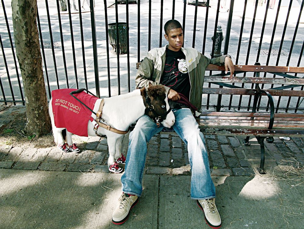 Man sitting on a city bench, with a miniature horse, wearing sneakers and a "Do Not Touch Service Animal On Duty" banner, standing beside him.