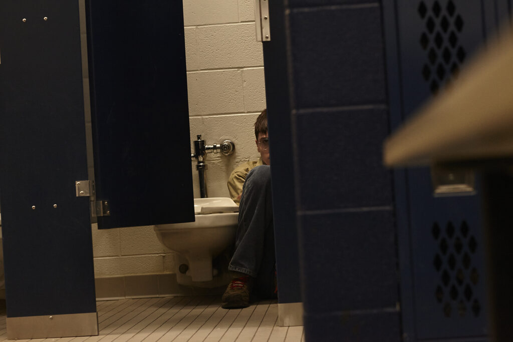 A young man with glasses sits on the floor of a bathroom stall