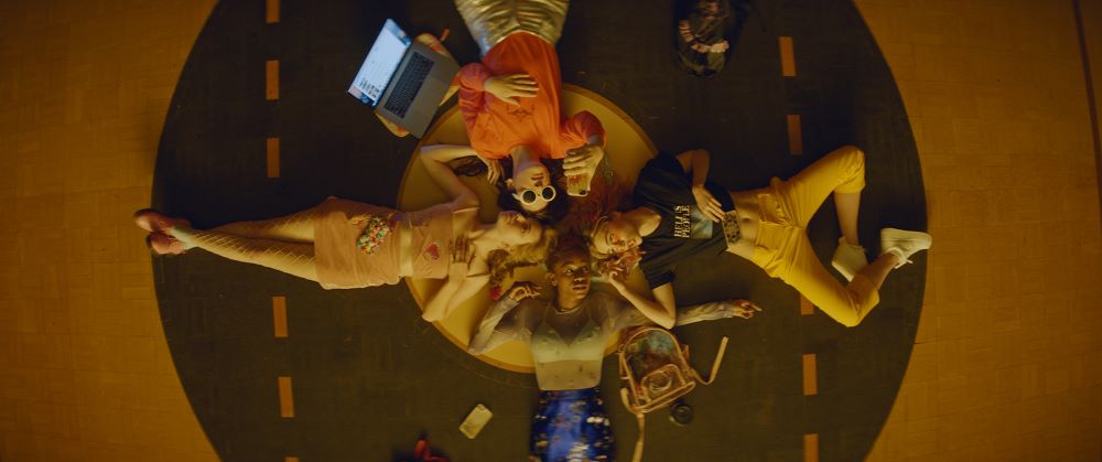 Four young women lie on their backs, head to head, holding smartphones and a laptop computer