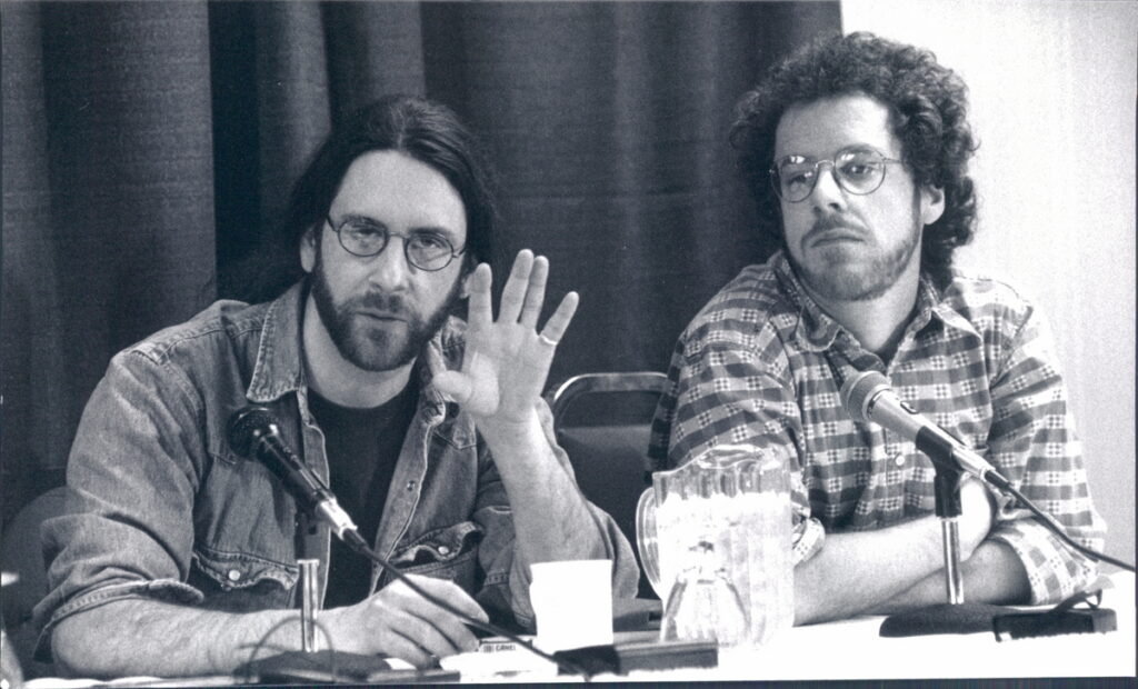 Two men sitting at a table, side-by-side, with microphones in front of them. The man on the observer's left has a dark beard and long, straight dark hair; he is wearing eyeglasses and a denim shirt; his left hand is raised at the elbow, as if he is gesturing while he speaks. The man on the right wears eyeglasses and a print shirt; he also wears a beard and has dark curly hair; his arms are folded in front of him.