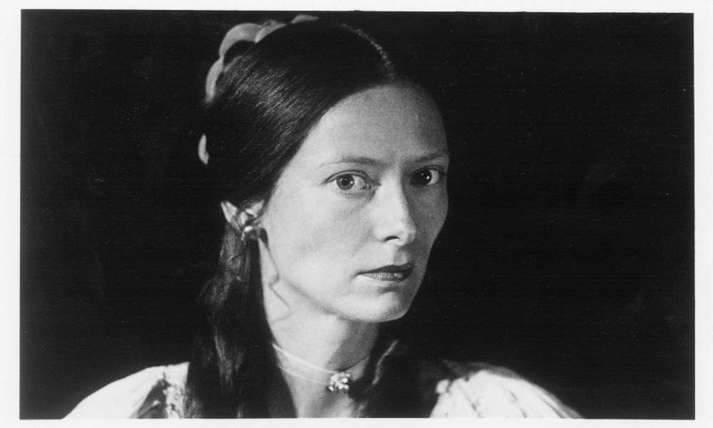 A woman with a 19th-century appearance gazes at the camera. She has long, dark hair and a choker-style necklace and serious eyes