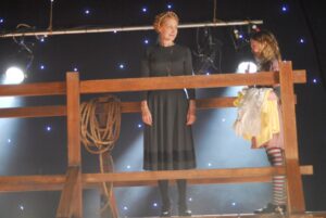 This appears to be a stage, with stage lights and artificial lights as stars in a night sky, above a sort of corral with a coil of rope hanging on one post. Inside is a woman in a black dress smiling at a young blond girl in a costume of some sort with red-and-white striped leggings.