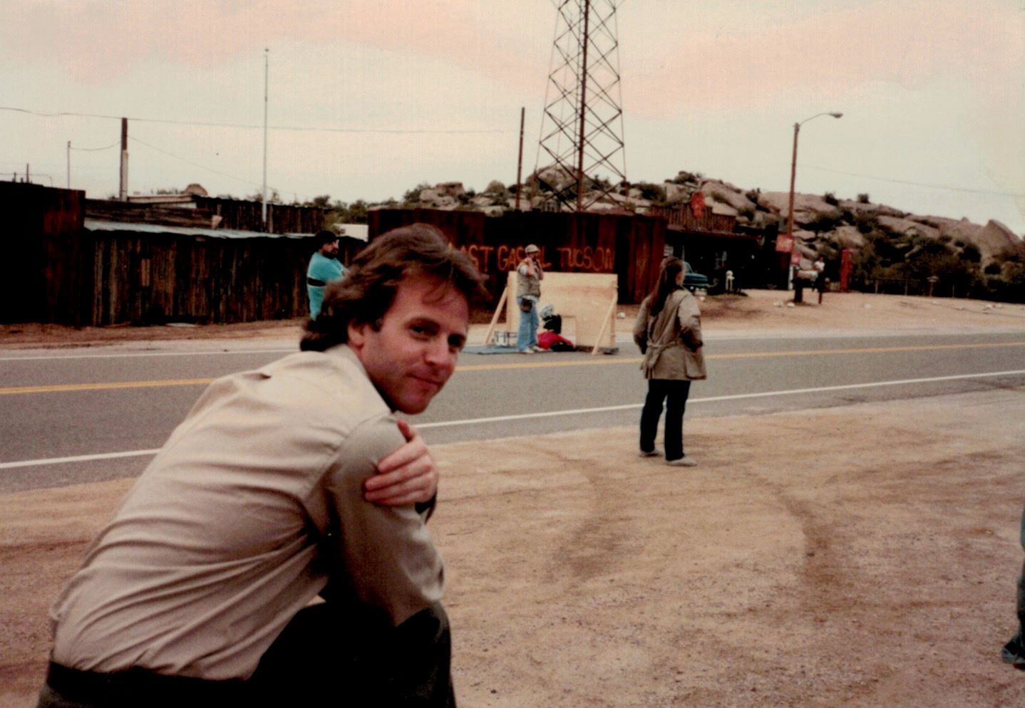 A man in a beige shirt and with medium-length dark hair kneels in the dirt and looks over his right shoulder at the camera