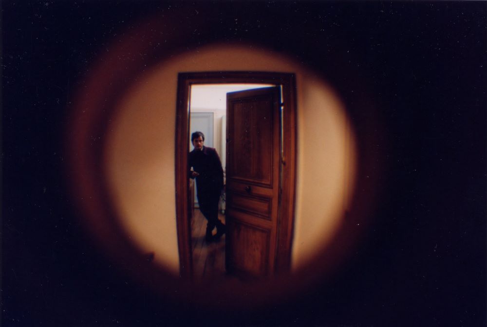 It's as if we're peering through a keyhold at the figure of a man standing in a room with an open door. He stands, leaning, legs crossed, peering at us.