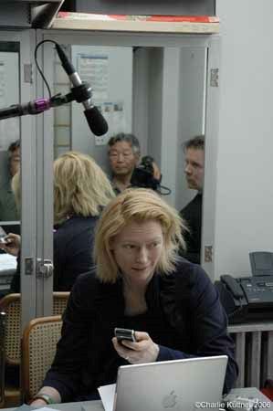 Blond woman smiles (worriedly) while sitting in an office setting with at least two men opposite her. One man appears to be holding a camera. The woman appears to be holding a cellphone or other small device. A Mac laptop is open before her.