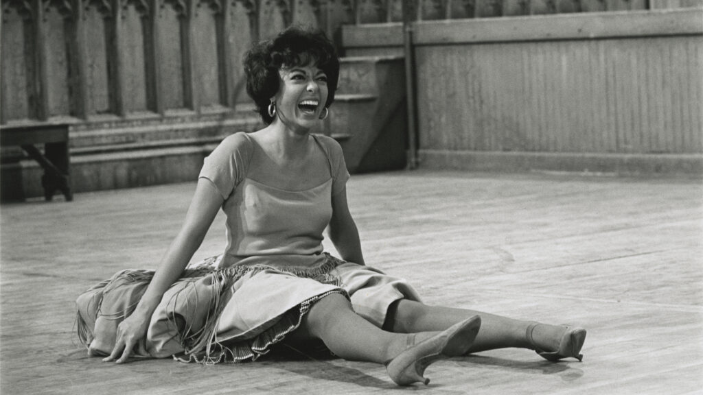 A black and white image of a young dancer sitting on the floor, smiling in a dress