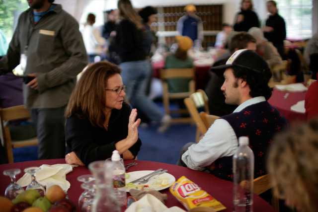 Woman and man sitting at a table in a busy lunchroom or restaurant. The brown-haired bespectacled woman is speaking and gesturing to the man, in a sweater vest and ball cap, facing her and listening.