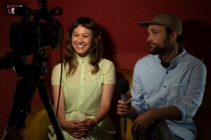 Smiling woman with long, light brown hair and bearded man in a ballcap sit in front of a movie camera pointed in their direction