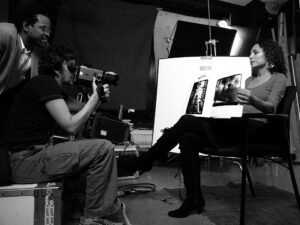 A man is pointing a camera at a woman holding a large white poster. Another man is standing close to him looking on.