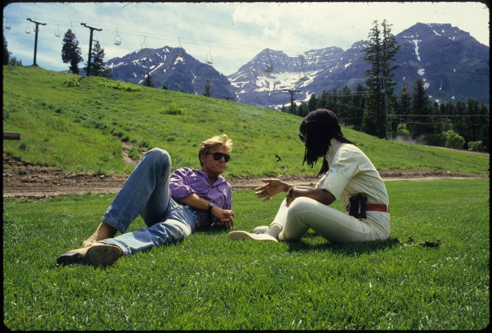 In a green field in a mountain setting, with a chair lift in the background, a blone man in sunglasses, jeans, light purple shoot reclines, speaking to a Black woman in white slcks and shirt, sitting cross-legged, face turned away from the camera