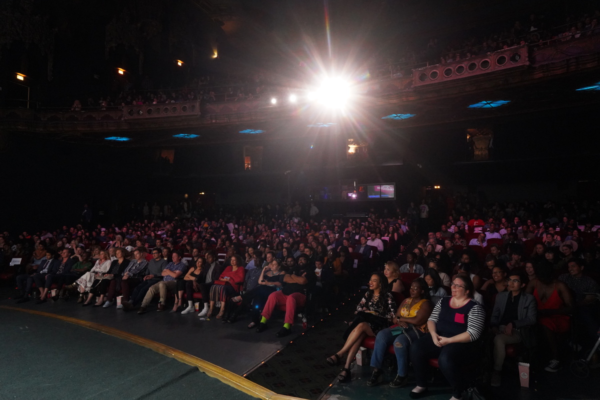 Audience members in a packed theater watching a movie. A bright projector light shines brightly.