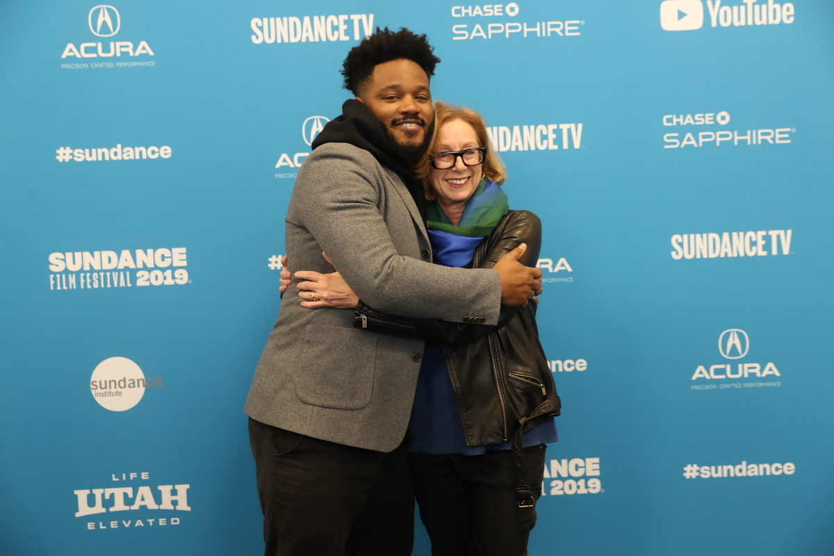 An image of a tall man (Ryan Coogler) in a grey jacket hugs a shorter woman (Michelle Satter) in a coat and blue scarf. The Sundance step and repeat is the backround