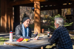 Ricardo speaks with Graham at a picnic table outside on a sunny morning and eating chips