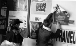 A man, with his back to the camera, hangs a poster with the words "Bottle Rocket" on a poster-laden wall. A woman with long brown hair in a ponytail, and a ball cap, holds a roll of tape.