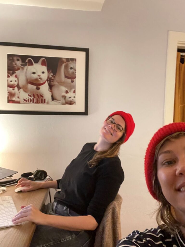 Woman with eyeglasses and long brown hair beneath a red stocking cap and another woman, with dark blond hair, also beneath a red stocking cap. only half of her face framed in the photo, work in what appears to be a home environment. A funk cat picture hangs on the wall.