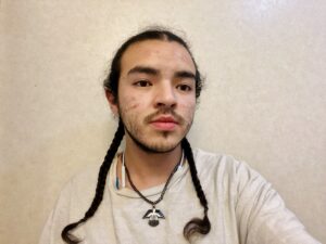 young man with two braids against a white wall looking at the camera