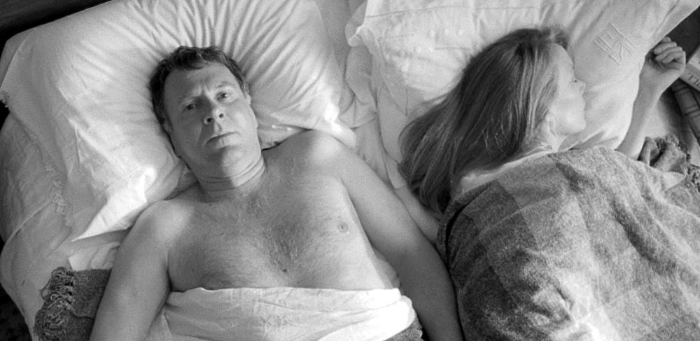 A middle-aged couple lie in bed, a bare-chested man lying on his back and staring at the ceiling on the left. On the right, a woman with long light brown hair lies on her side, seemingly asleep, facing away from the man,