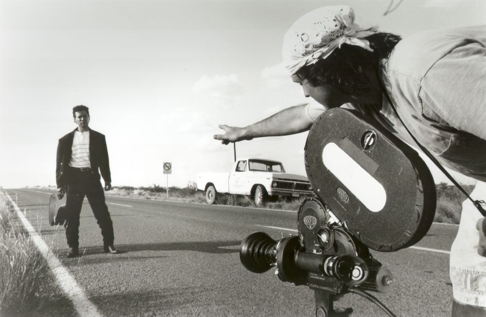 Young man with bandanna tied on head stands, bent at waist, over a camera stationed on a highway, right arm extended as if to direct another man. standing several feet down the highway. The second man wears a dark jacket and slacks, a light shirt with horizontal stripes. He carries a guitar case in his right hand. In the background, a light-colored Ford pickup is parked off the side of the highway.