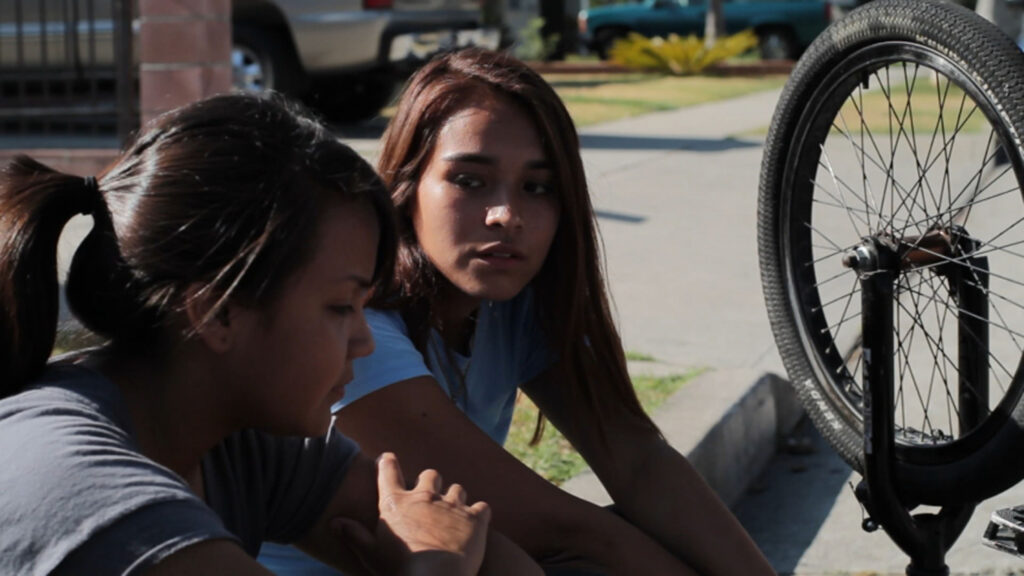 two young women sitting on a curb looking at each other with an upside down bicycle