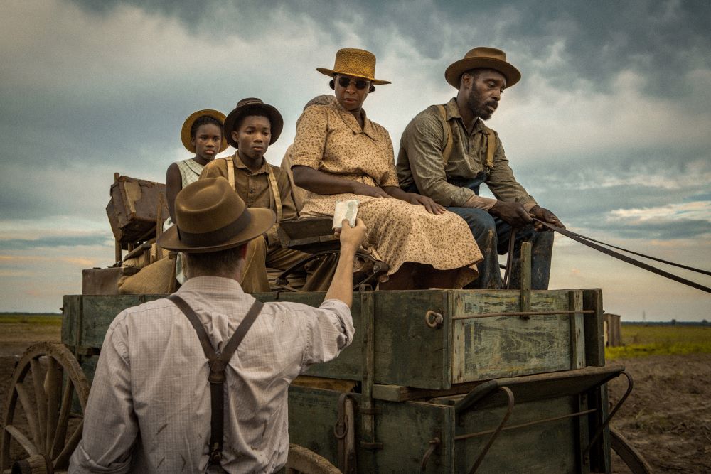 A Black family rides in a horse-drawn wagon, man and woman up front, boy and girl in back. A white man, with back to the camera, is handing what appears to be a paper to the woman on the near side.