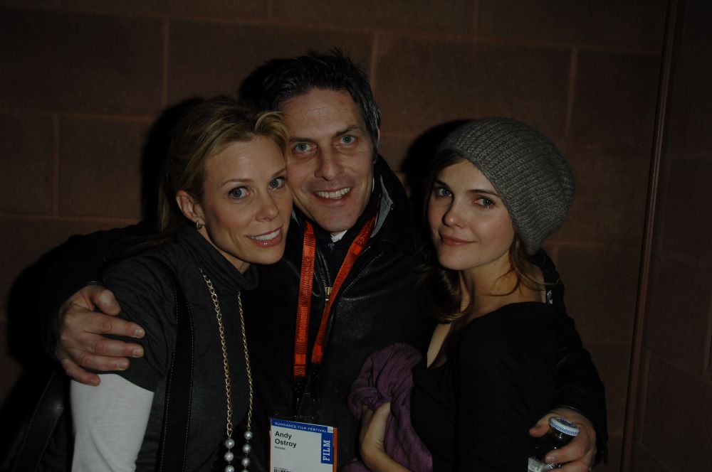 A man with hair graying at the temples and a lanyard printed with "Andy Ostroy" hugs two women on each side of him. On the left is a smiling blondish woman, and on the right is a woman with brownish hair under a gray stocking cap, also smiling slightly.