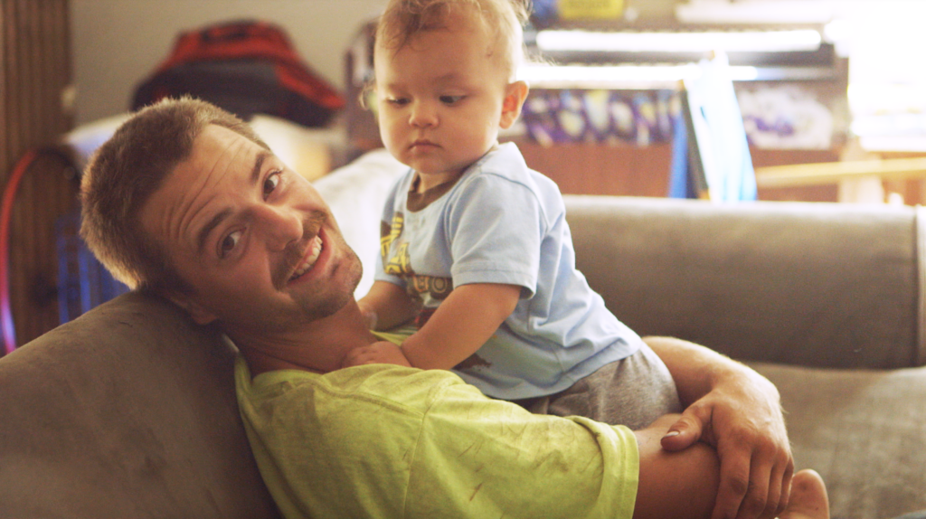 Man in his 20s holding a baby smiling at the camera while sitting on the couch