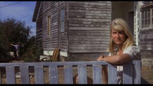 Blonde woman off to the right looking at the camera out on a porch in front of a house