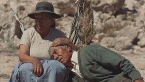The background is an arid environment. An older Indigenous woman, in a brown fedora-type hat, beige top and blue skirt, sits, cradling the head of an older Indigenous man, dressed in a light-colored collared shirt and green cardigan sweater. Both gaze at something to their right.
