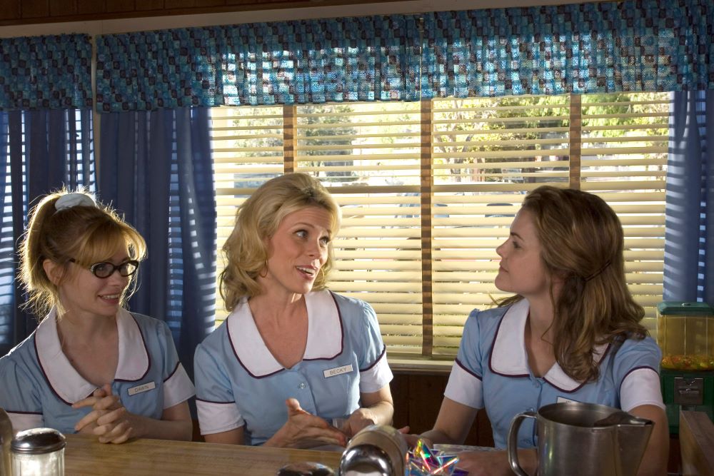 Three women in blue waitress uniforms with white collars sitting side-by-side at a table or counter and in front of a window, showing daytime light through blinds. From left, a slightly smiling woman with dark-framed eyeglasses, with blond hair swept up in what appears to be a high ponytail and the nametag "Dawn"; another blond woman apparently speaking to the third woman, smiling and animated, nametag "Becky"; a woman with long, light brown hair, chin up as she listens to Becky, nametag is blocked by a metal water pitcher.