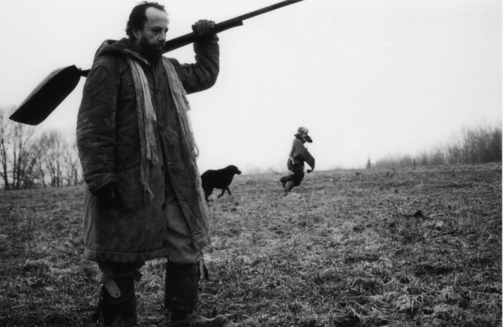 Bearded man carrying a shovel over his shoulder. What appears to be a child and a dog are running in the background. The photo is black and white.