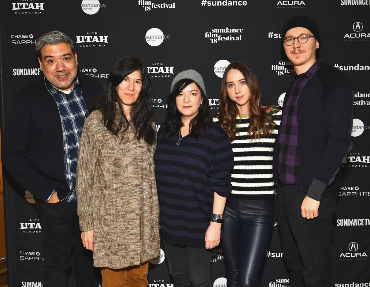 A man in a black jacket and blue triped shirt, a woman in borwn with long black hair, a woman with a striped black and white shirt and long brown hair, and a man in black with glasses and a black beanie stand in front of the 2018 Sundance Film Festival step and repeat.